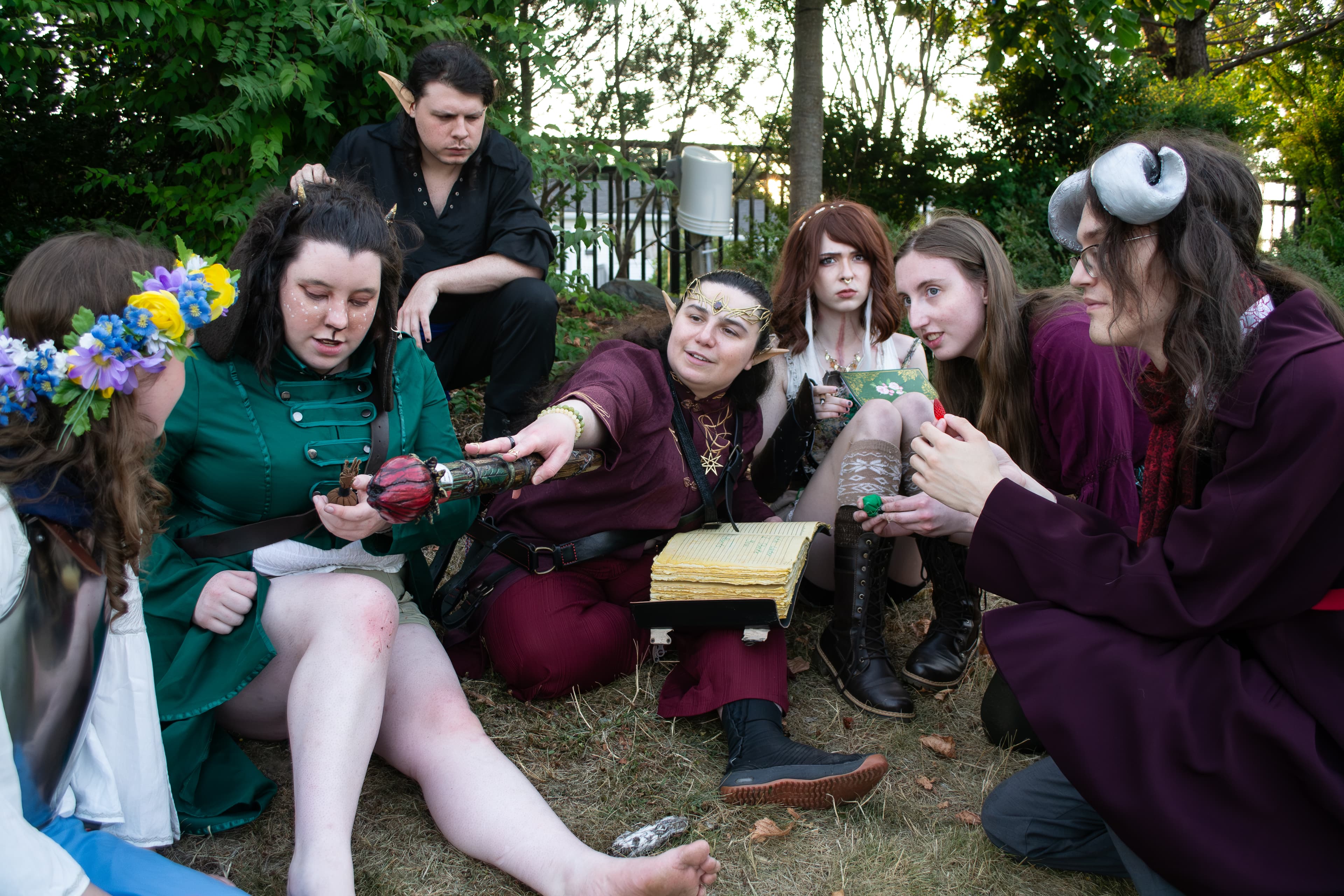 Seven people wearing fantasy costumes sitting on the ground. One person has an open book on their lap and is pointing a wand at another.
