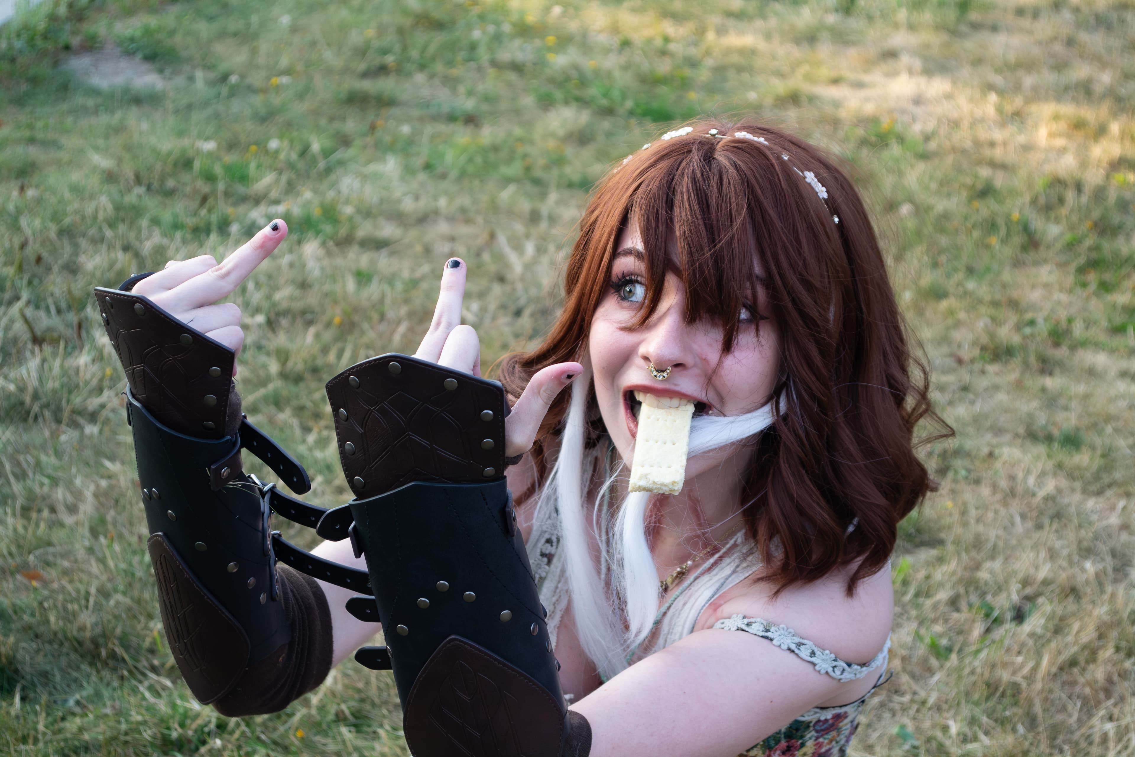A person wearing a fantasy costume with disheveled hair and a piece of shortbread between their teeth sticks their middle fingers up at someone off-camera.