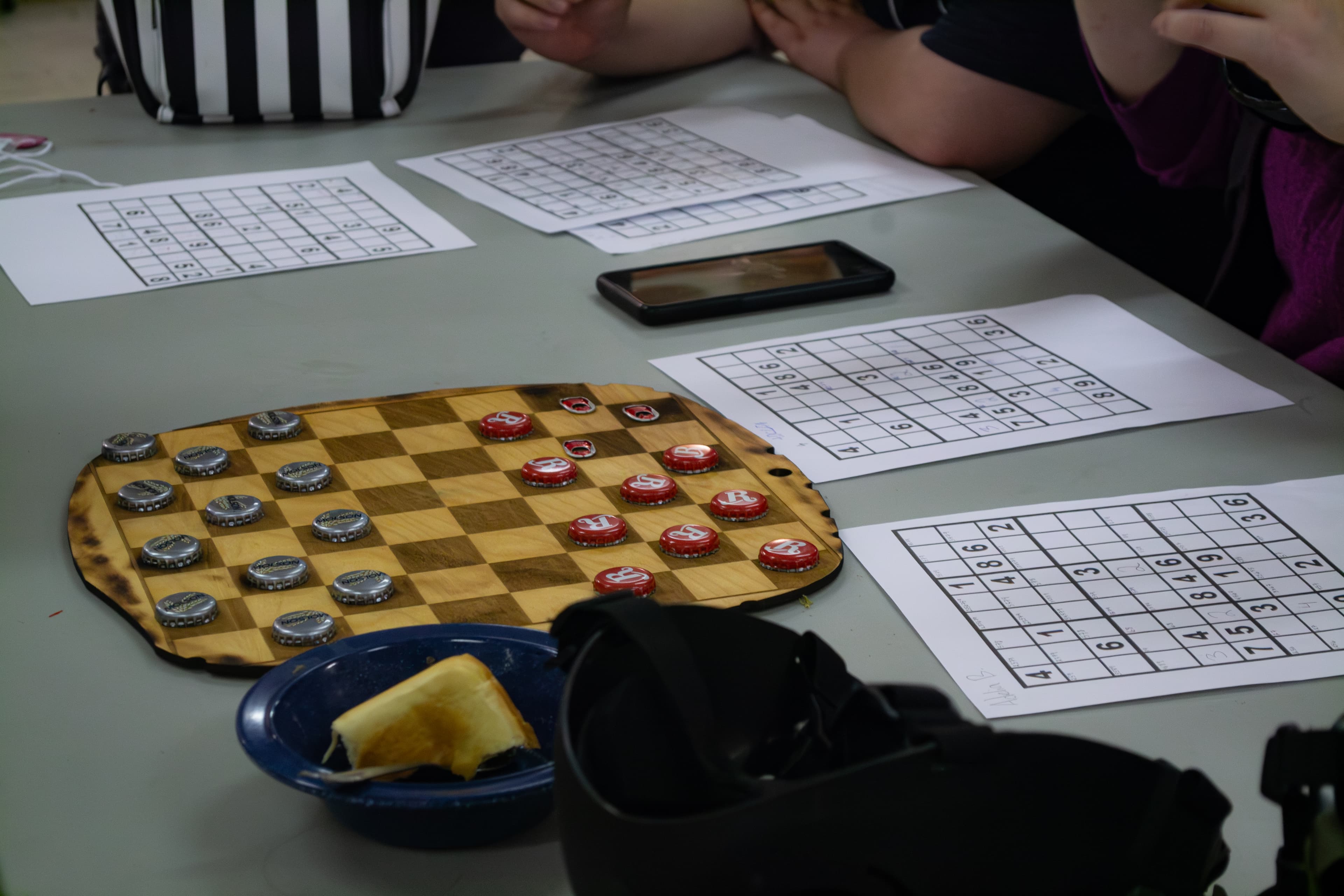 A table covered in miscellaneous items. The focus is on a checkerboard with bottle cap playing pieces, and five sudoku sheets people are working on.