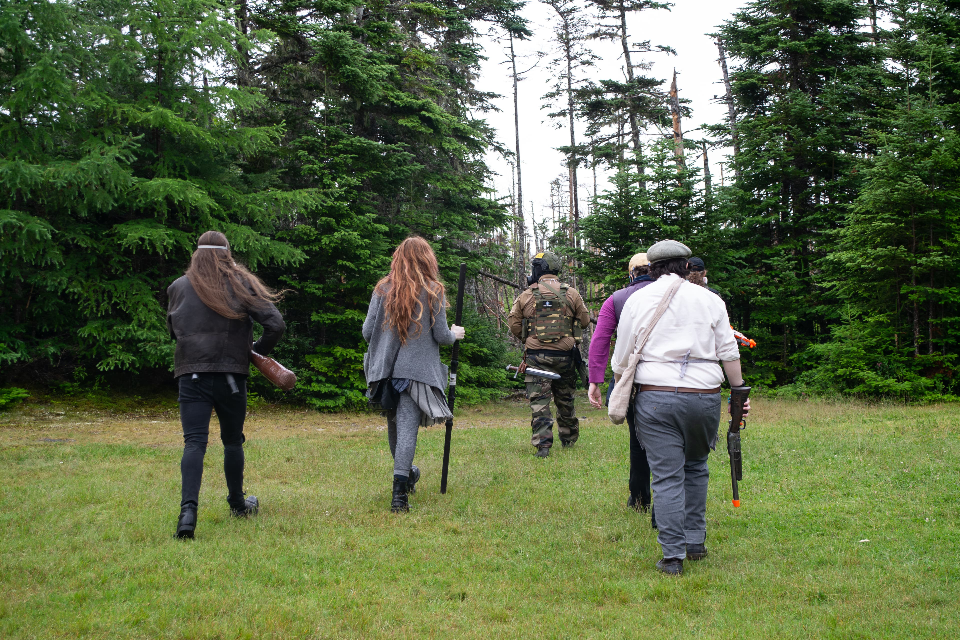 Six people wearing post-apocalyptic costumes and carrying prop weapons walking towards a forest with their backs facing the camera.