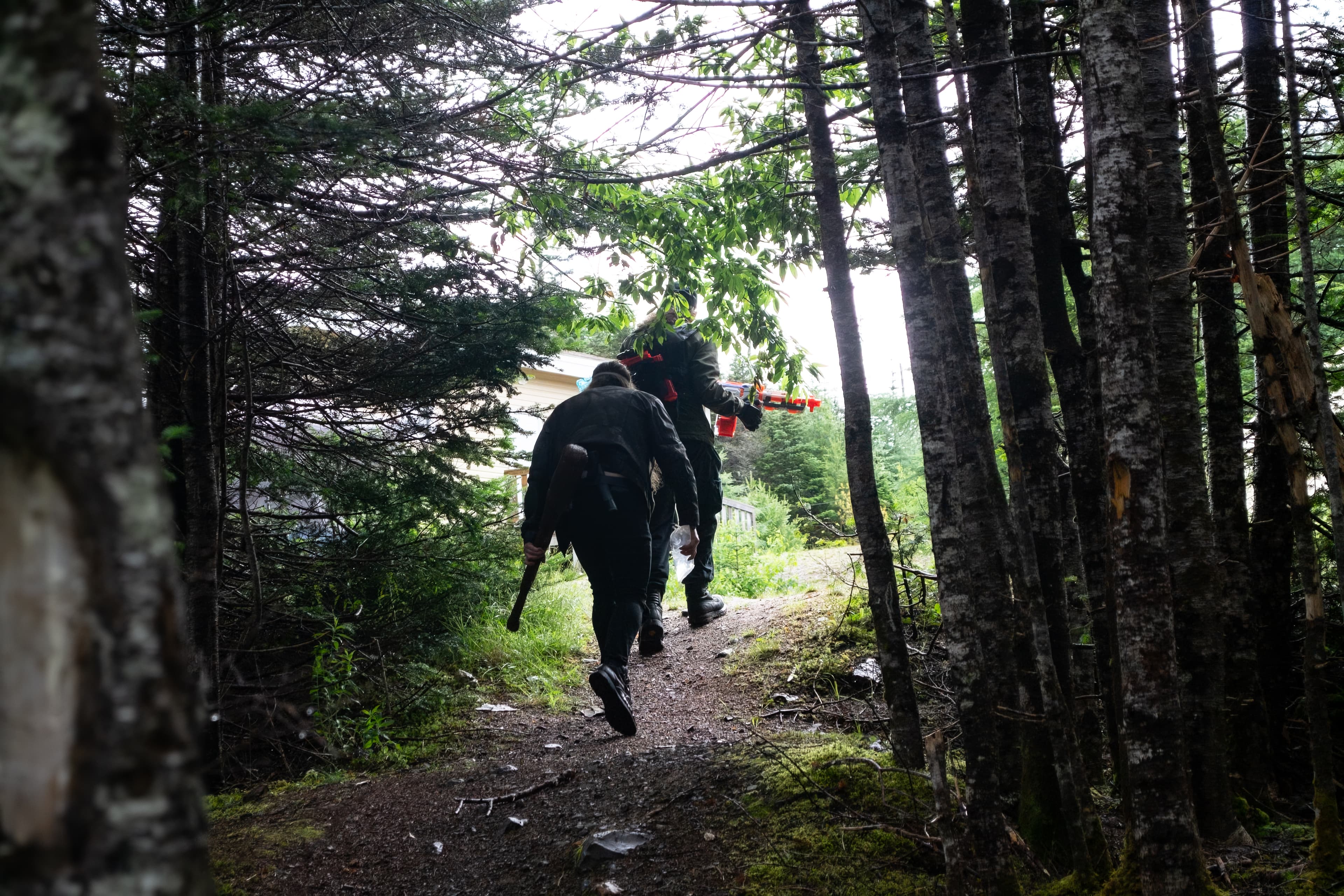 Two people wearing post-apocalyptic costumes and carrying prop weapons walking on a path exiting a forest with their backs facing the camera.