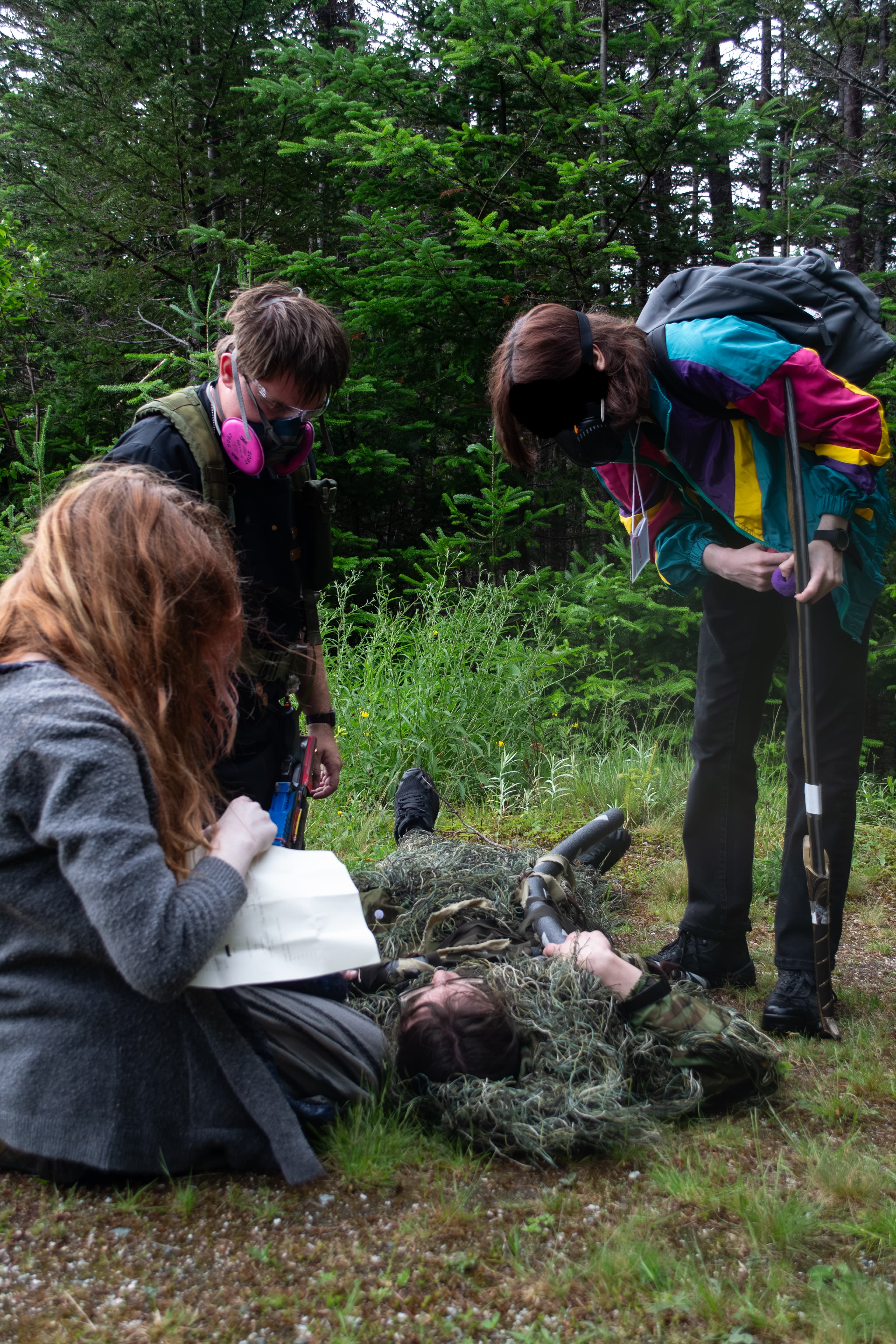 Three people wearing respirators looking down to examine another person laying on the ground wearing a ghillie suit.